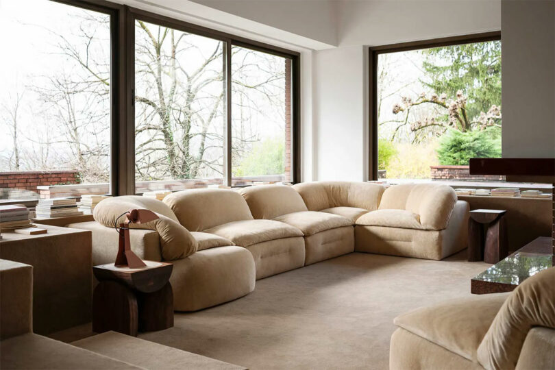 Spacious living room with a large beige sectional sofa, low tables, books, and floor-to-ceiling windows showing trees and greenery outside, accented by Faye Toogood-inspired design elements.