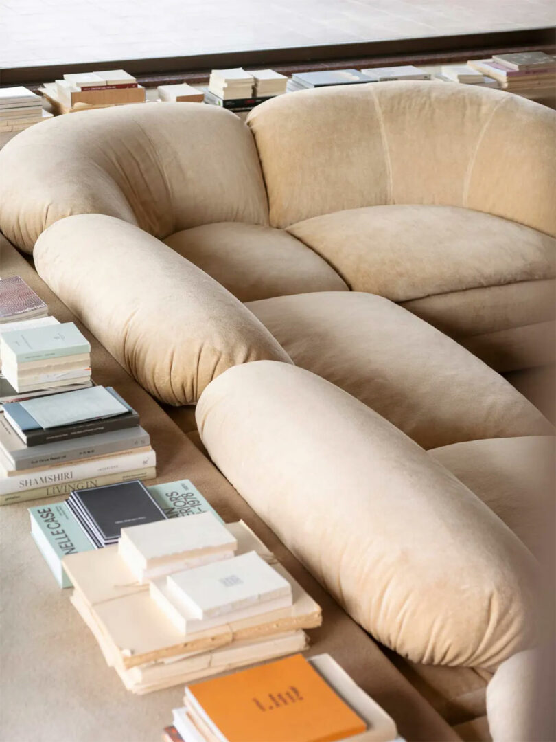 Curved beige velvet Faye Toogood sofa surrounded by stacks of books on a low coffee table in a modern, minimalist living space.