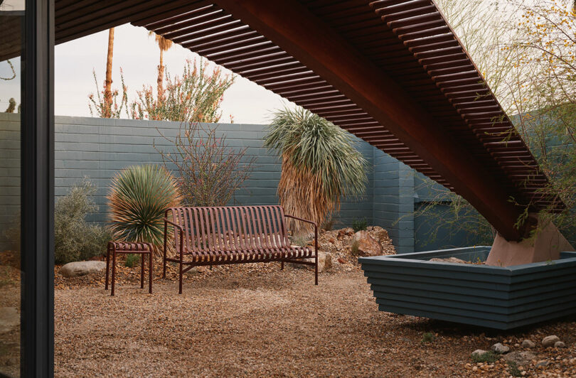A metal bench sits on gravel under a slatted wooden structure in a modern, enclosed garden with desert plants and a painted brick wall.