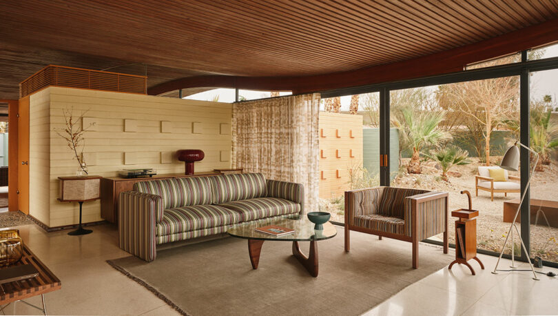 Mid-century modern living room with striped sofa, wooden armchair, glass coffee table, and floor-to-ceiling windows overlooking a desert garden.
