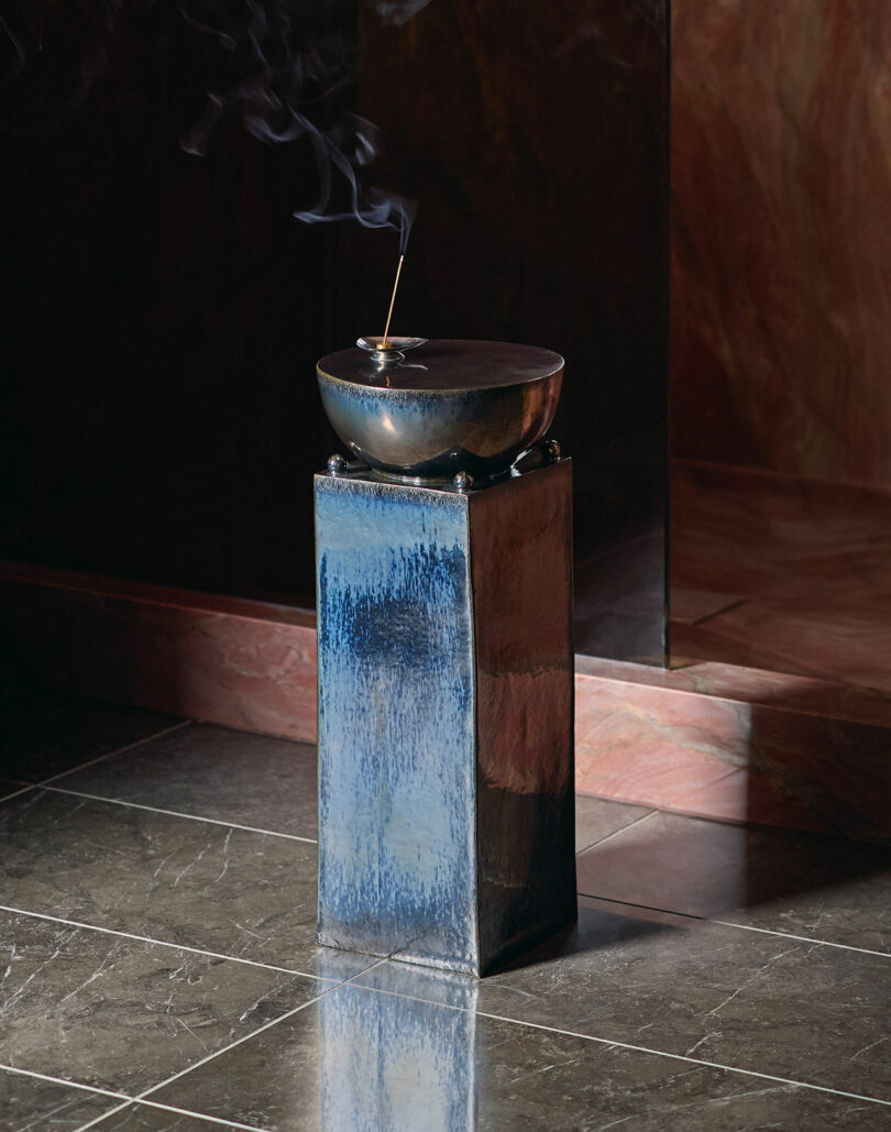 A metal bowl with a lit incense stick, emitting smoke, sits atop a blue ceramic pedestal on a tiled floor.