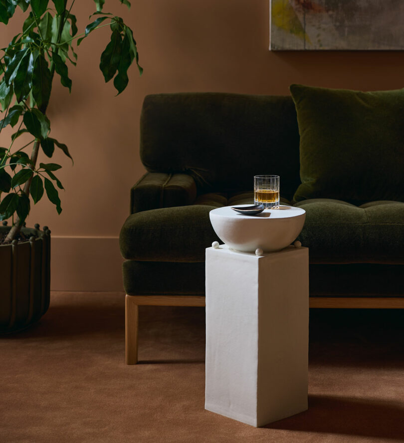 A white pedestal with a round bowl, spoon, and glass sits in front of a dark green sofa; a potted plant and abstract art are in the background.