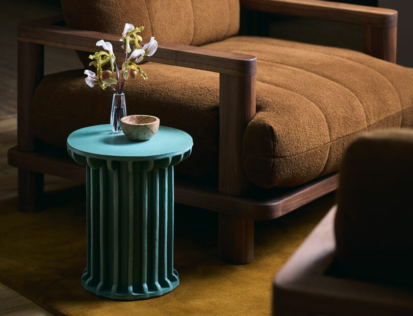 A green ridged side table holds a small glass vase with white flowers and a ceramic bowl next to a brown cushioned armchair.