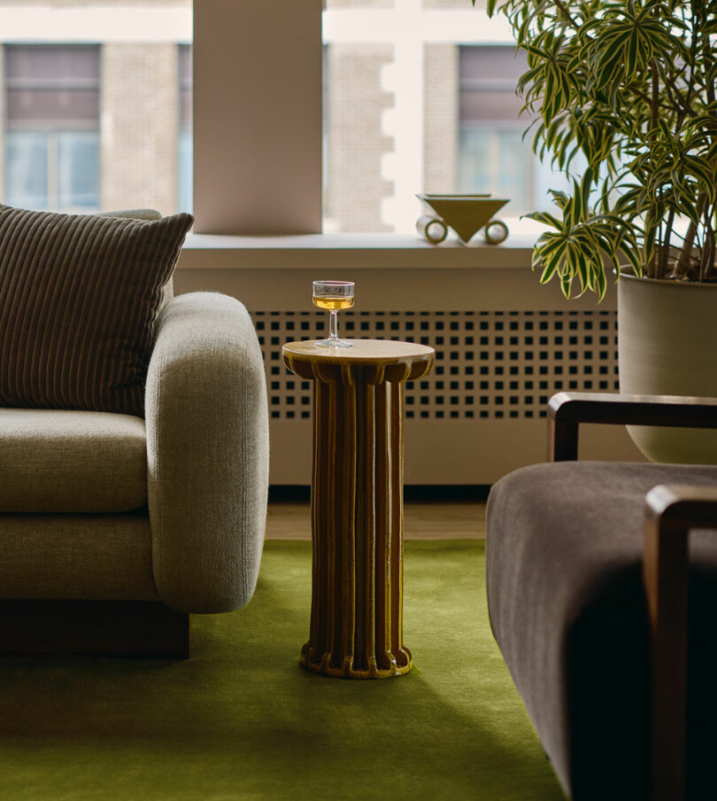 A glass of drink sits on a small gold side table between a light gray sofa and a dark brown armchair in a modern living room with green carpet and a potted plant.