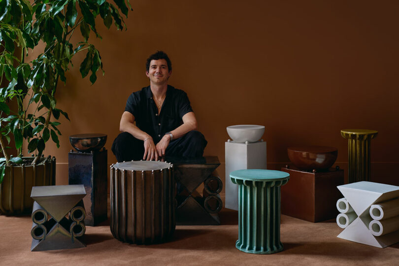 A person sits surrounded by various modern, geometric stools and tables in a room with brown walls and a large green plant.