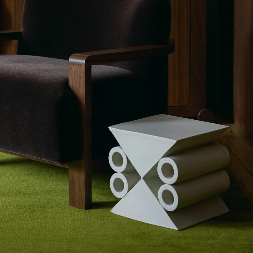 A modern geometric side table with cylindrical and angular shapes sits next to a dark armchair on a green carpet.