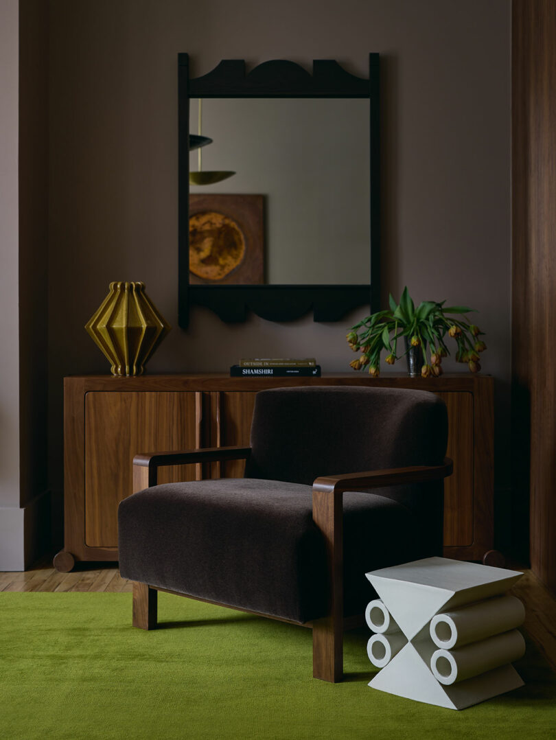 A dark armchair sits on a green rug in front of a wooden cabinet, with a mirror, lamp, plant, and books arranged as decor. A sculptural white side table holds rolled magazines.