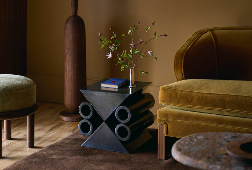 A modern living room features a sculptural side table with a vase of flowers and a closed book, next to a mustard yellow velvet sofa and other wooden furnishings.