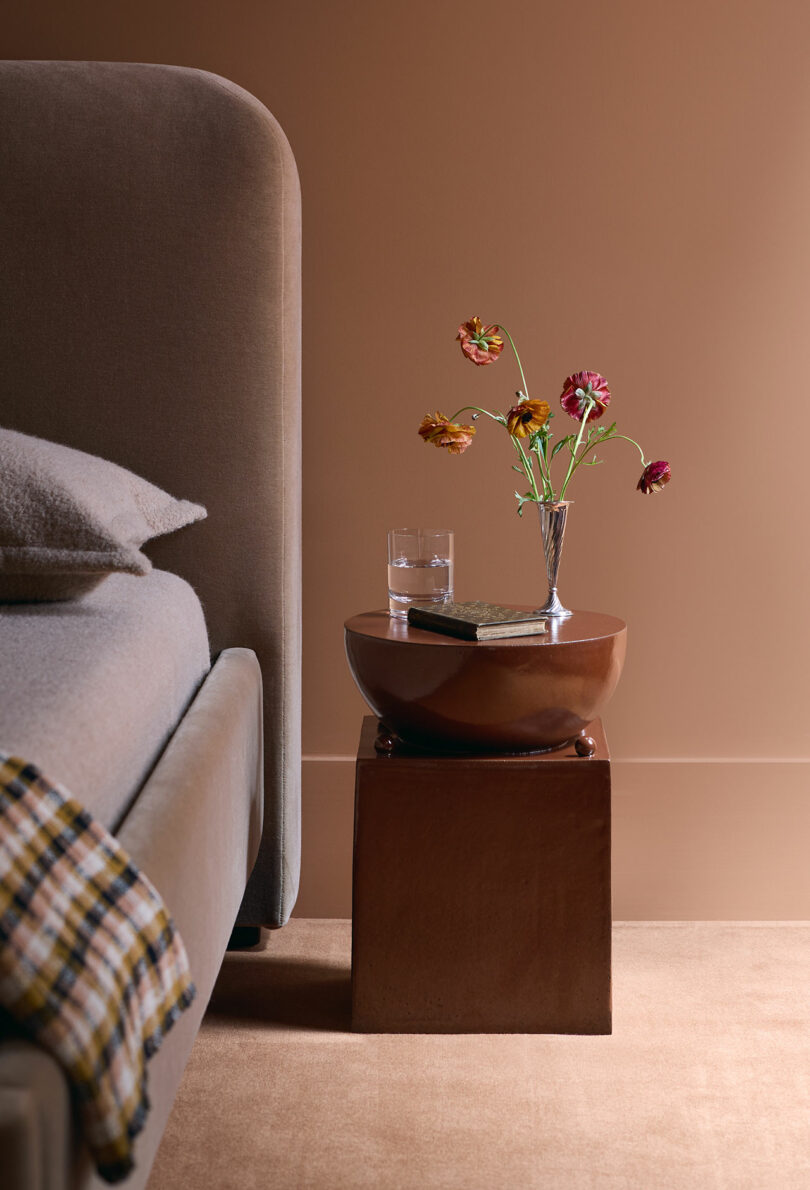 A modern bedroom scene with a taupe bed, a brown nightstand holding a glass of water, a small book, and a vase with colorful flowers against a beige wall.