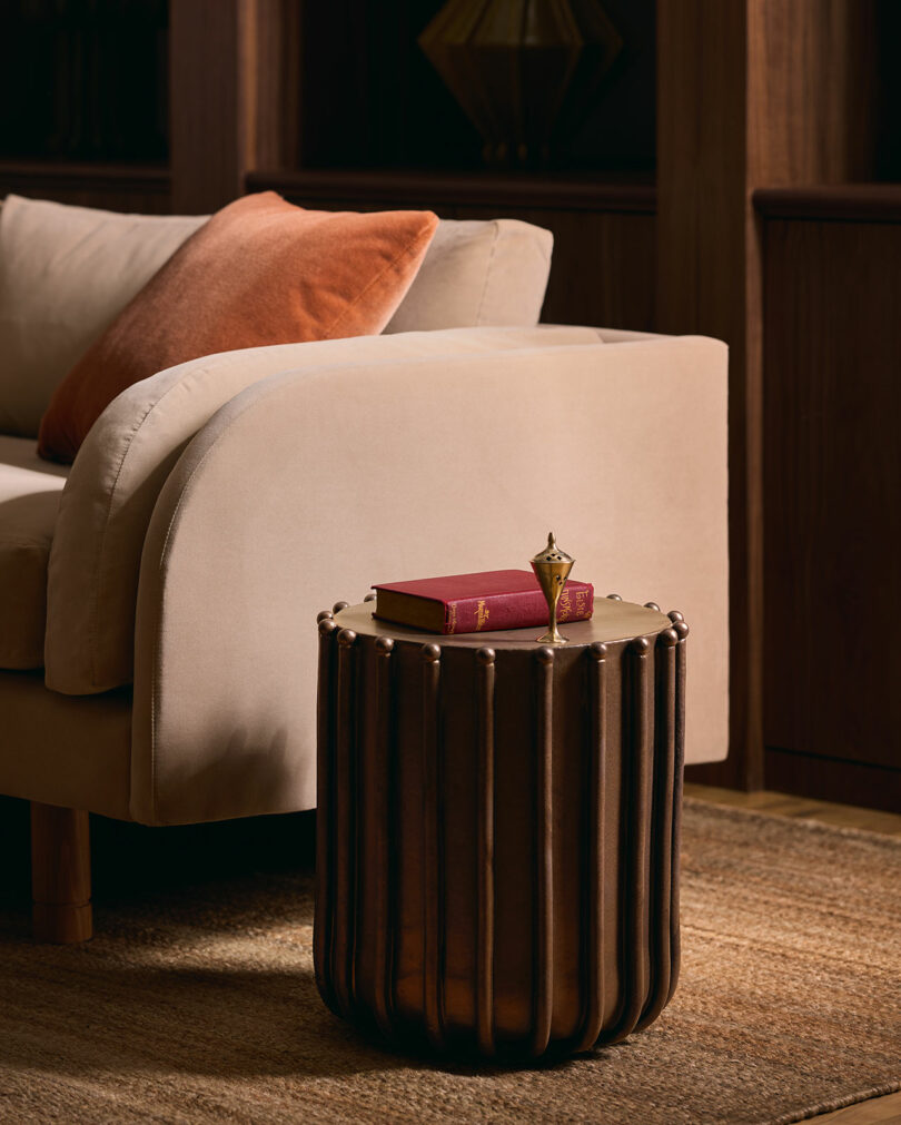 A small bronze side table holds a red book and a decorative object next to a beige sofa with an orange cushion on a woven rug.