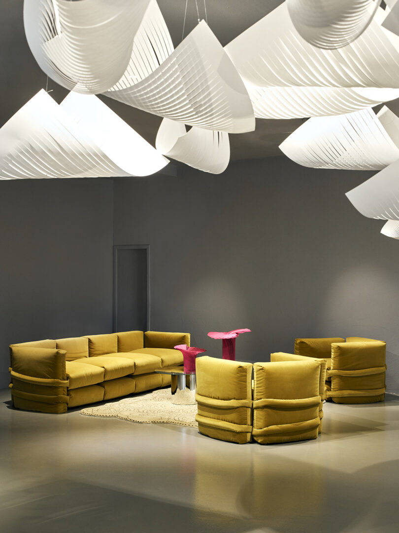 A modern lounge with mustard yellow sectional sofa and chairs, a pink side table, a beige rug, and sculptural white ceiling lights in a minimalist gray room.