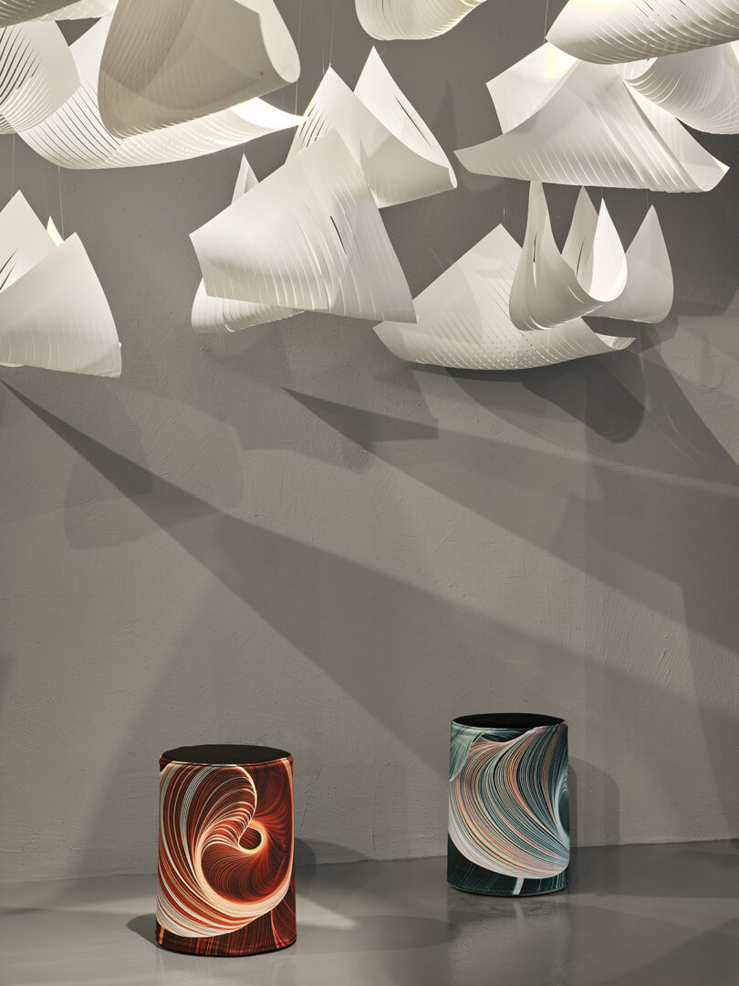 Two cylindrical stools with swirling patterns sit beneath suspended, sculptural white paper forms casting shadows on a plain gray wall.