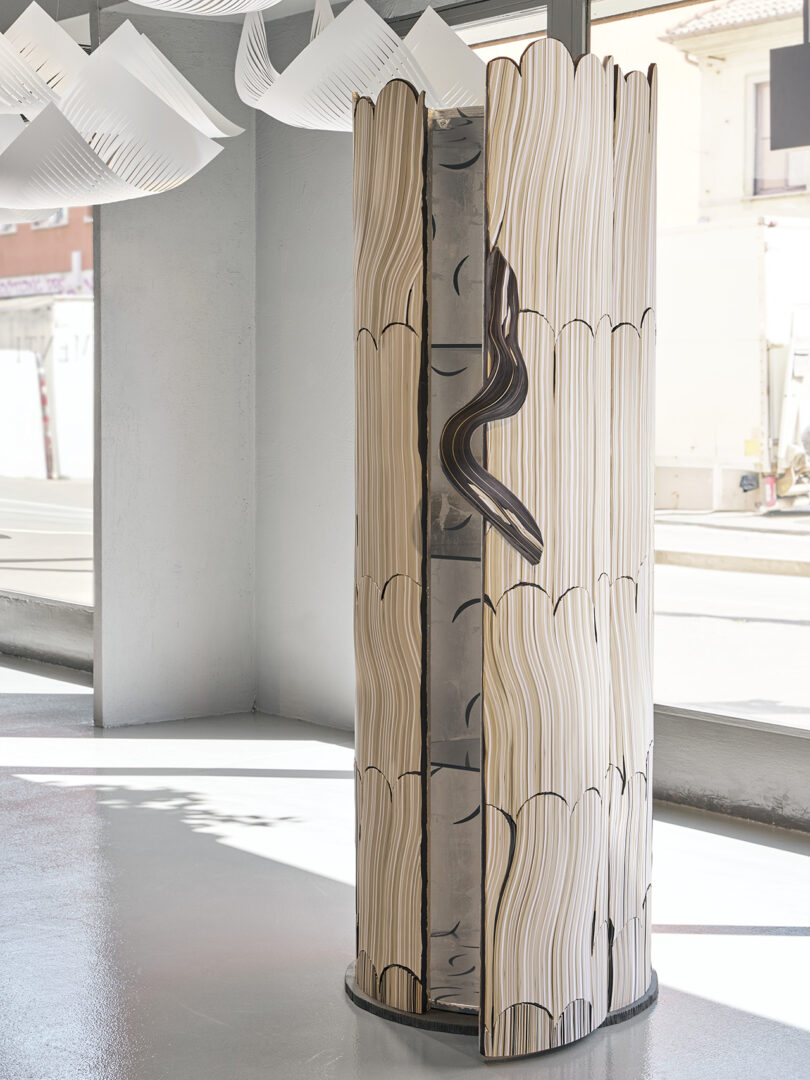 A freestanding sculptural screen resembling stacked, wavy book pages with a snake-like form entwined around its center, displayed in a bright, modern gallery space.