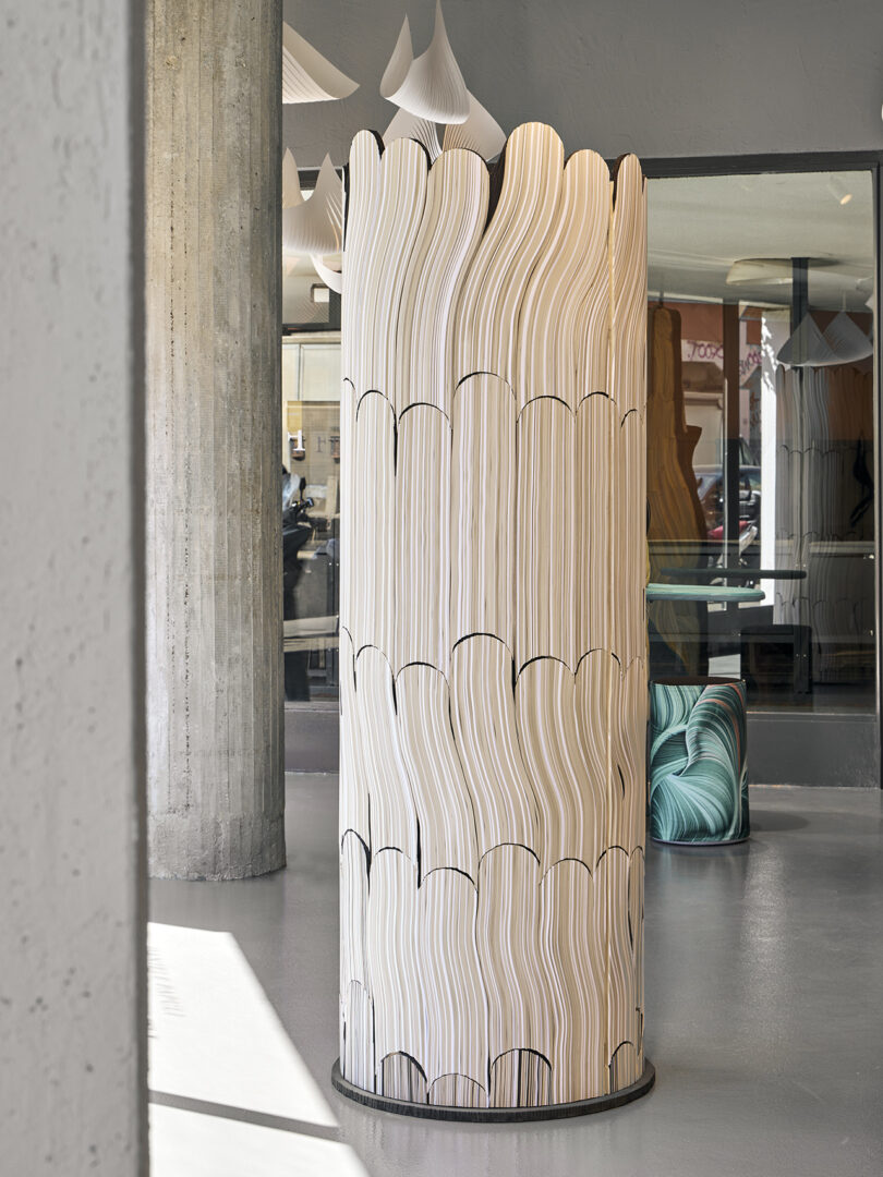 A freestanding, cylindrical partition made of wavy, layered wooden panels sits on a polished concrete floor in a modern interior space.