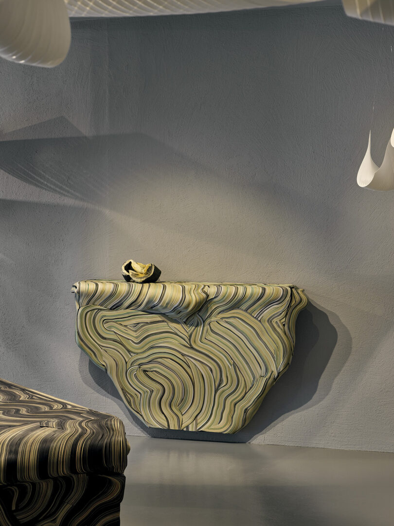 A sculptural console table with curved, wavy marbled patterns is mounted against a textured gray wall; a small matching bowl sits on top.