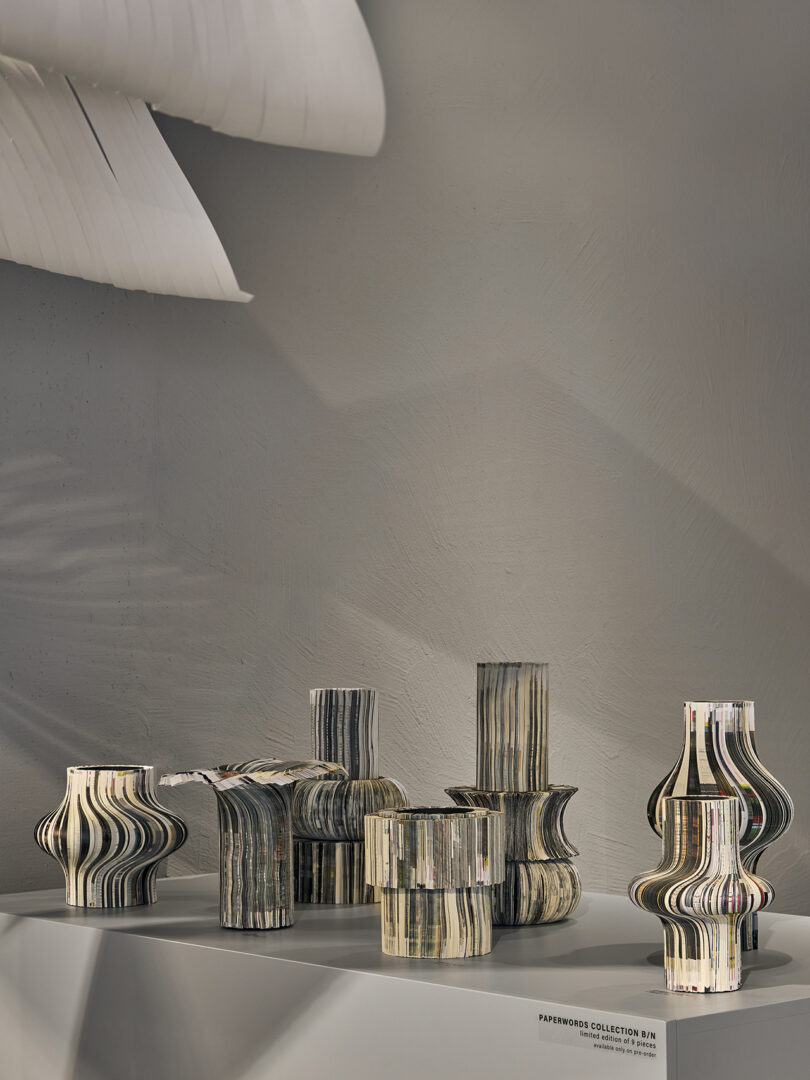 A group of sculptural vases made from layered, recycled paper is displayed on a white platform against a neutral wall.