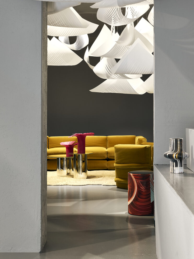 A modern living room with a mustard yellow sectional sofa, metallic tables with pink accents, a white rug, abstract ceiling lights, and sculptural decor pieces.