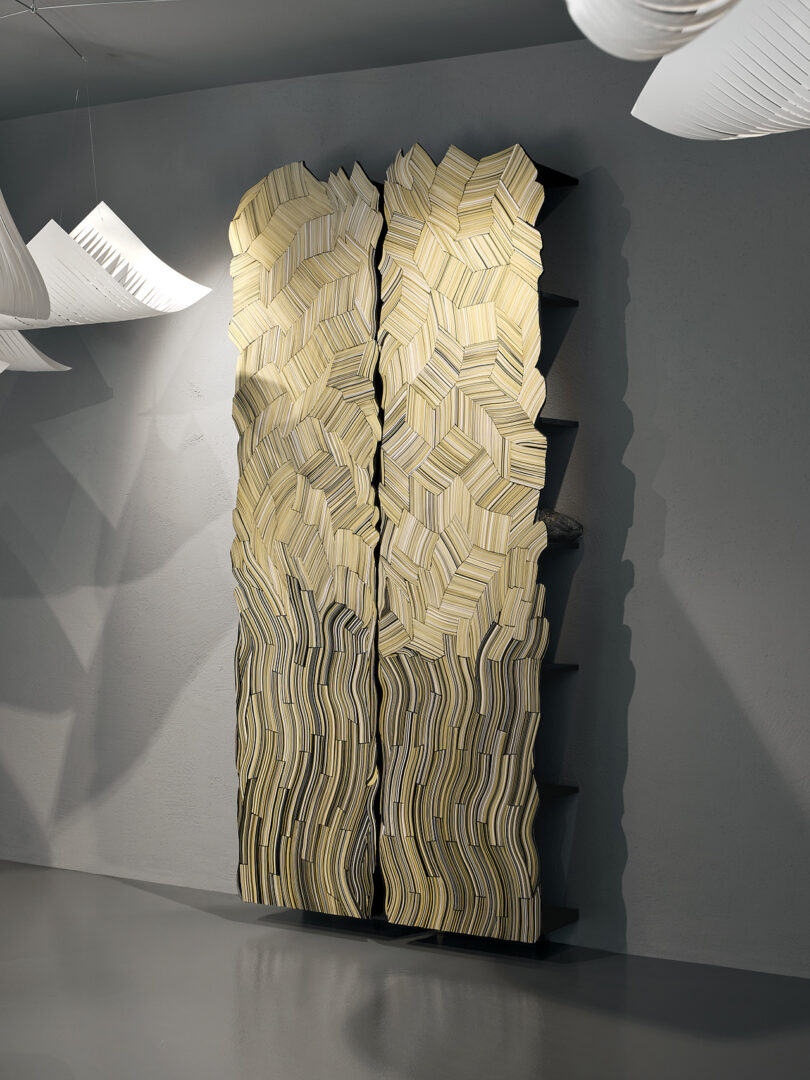 A sculptural bookshelf with staggered, overlapping beige book-like forms creating a textured geometric pattern on a gray wall.