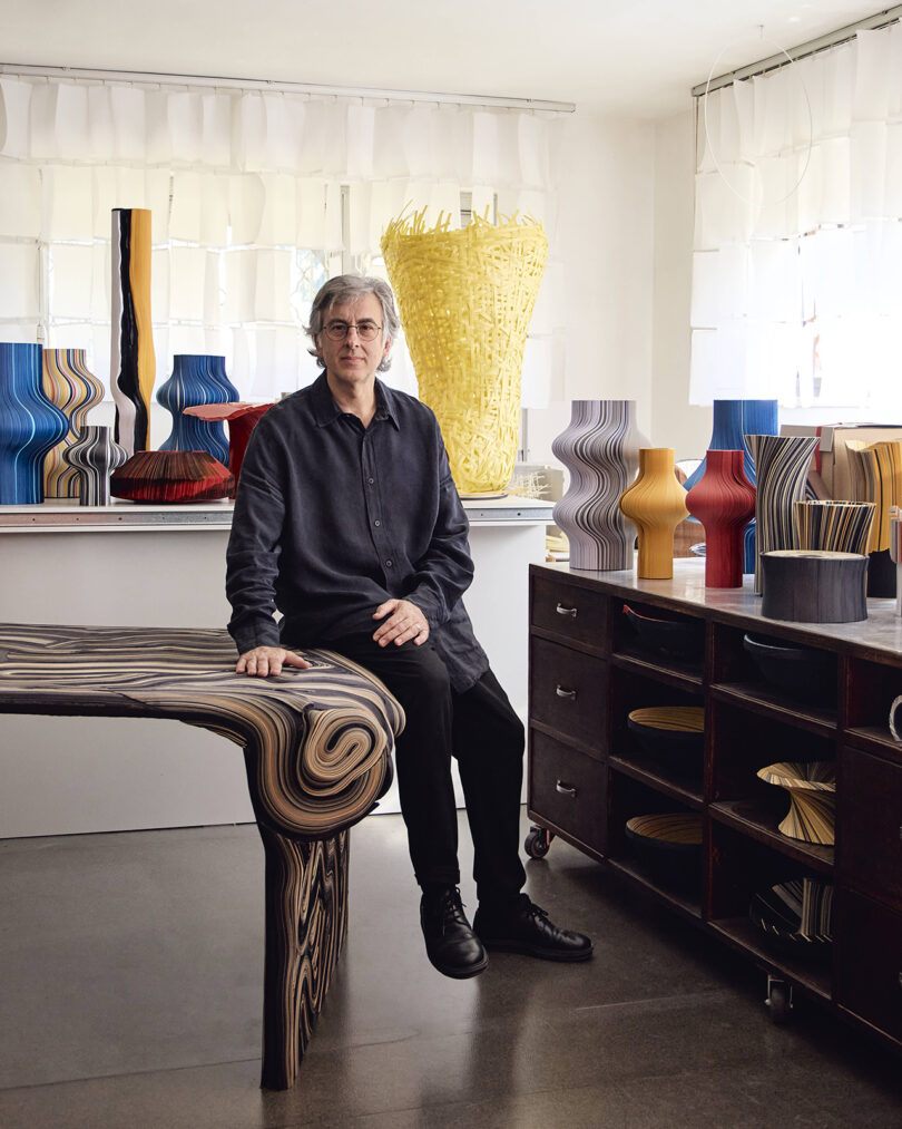 A person sits on a sculptural table in a studio filled with colorful, patterned vases and vessels, with shelves and counters displaying more objects in the background.