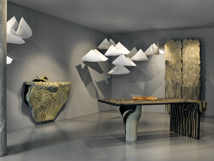 Modern room with sculptural wood-grain patterned furniture, including a table, console, and screen, with hanging white geometric paper light fixtures on a gray floor and walls.
