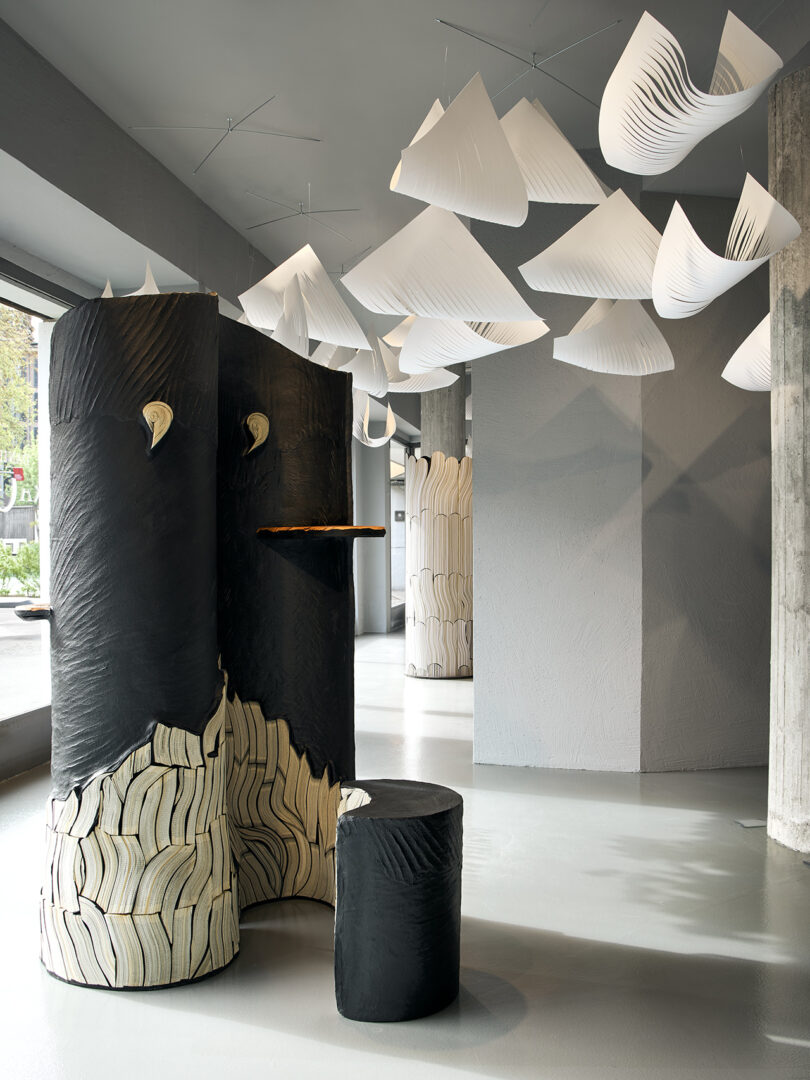 Modern interior with sculptural black and white columns, a round stool, and hanging geometric white paper-like shapes from the ceiling. Natural light enters through large windows.