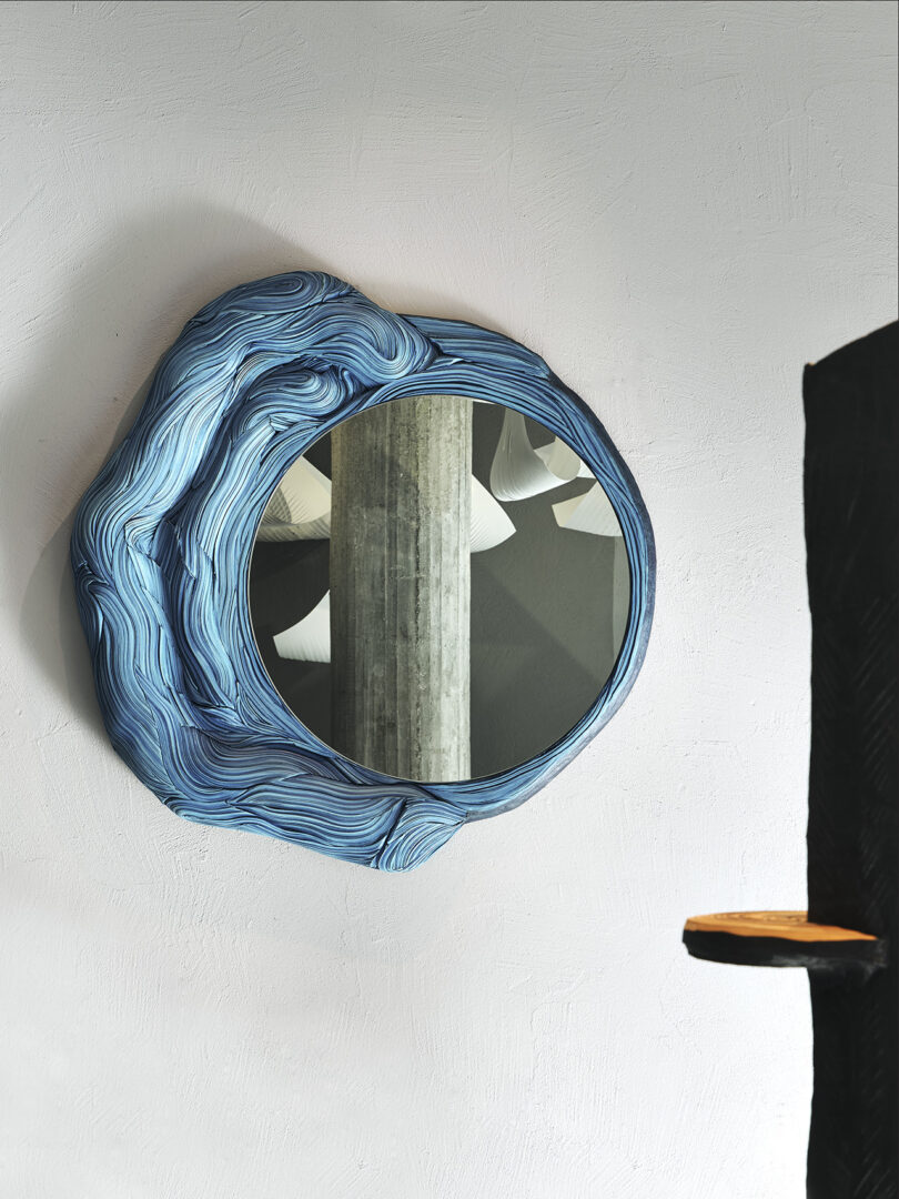 A round mirror with a textured, wavy blue frame hangs on a light-colored wall, reflecting a pillar and ceiling lights. Part of a dark object and a small wooden shelf are visible on the right.