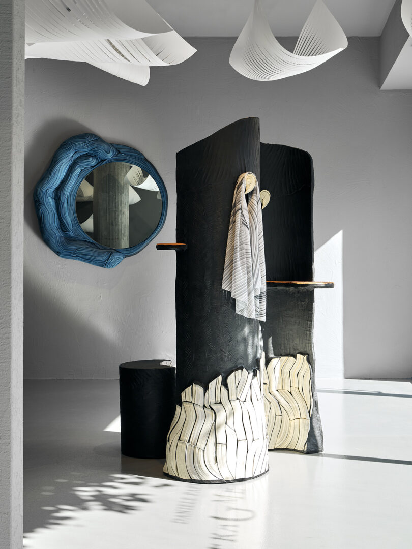 A modern room with sculptural black and white divider panels, a round blue mirror on the wall, and draped striped fabric, with soft natural light and white ceiling decorations.