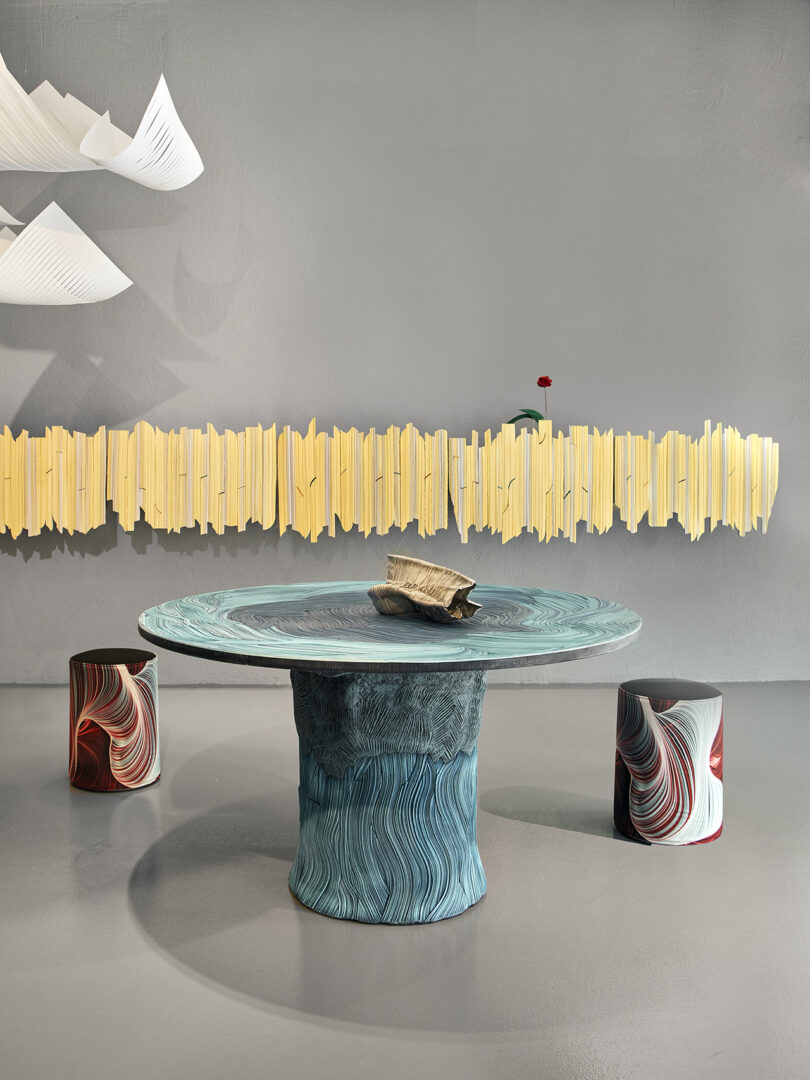 A round, blue textured table with two patterned stools sits in front of a gray wall, featuring a yellow sculptural wall piece and a white hanging light fixture above.