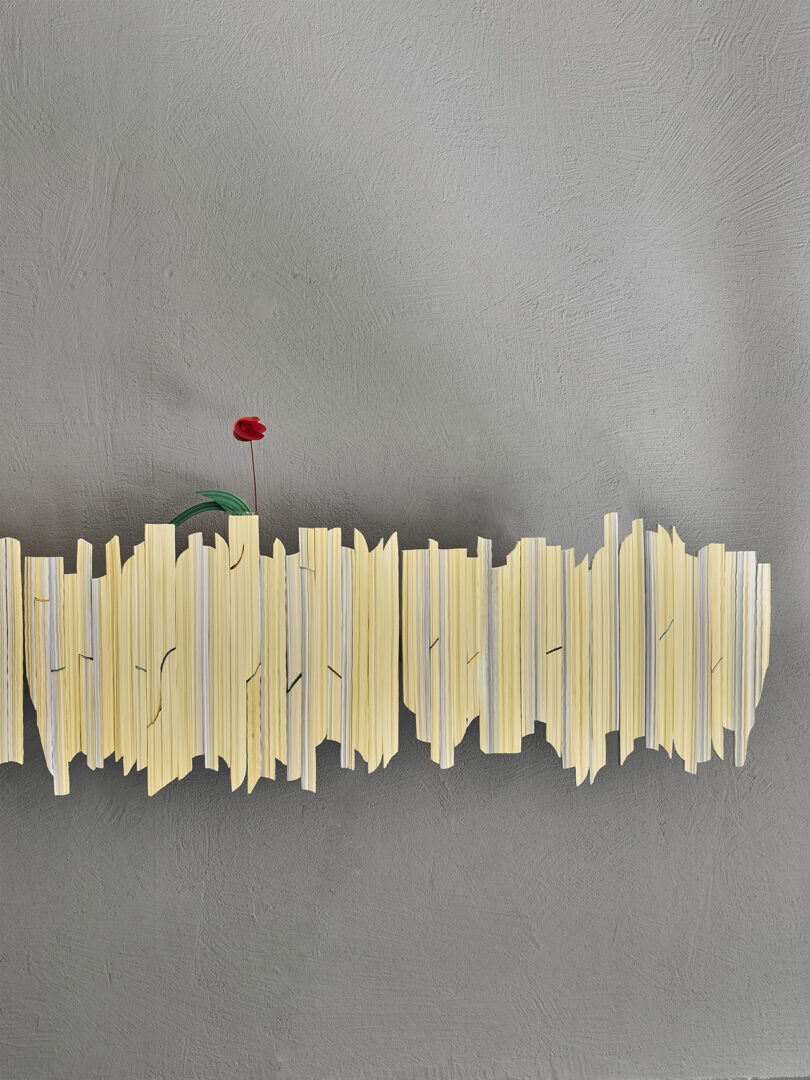 A single red flower in a green vase is placed on a jagged, abstract yellow shelf against a textured gray wall.