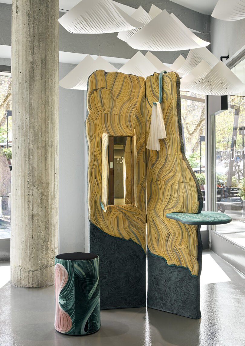 Sculptural installation with textured yellow and green panels, a small round table, a mirror, and a matching green and pink round stool, set in a modern, light-filled space.
