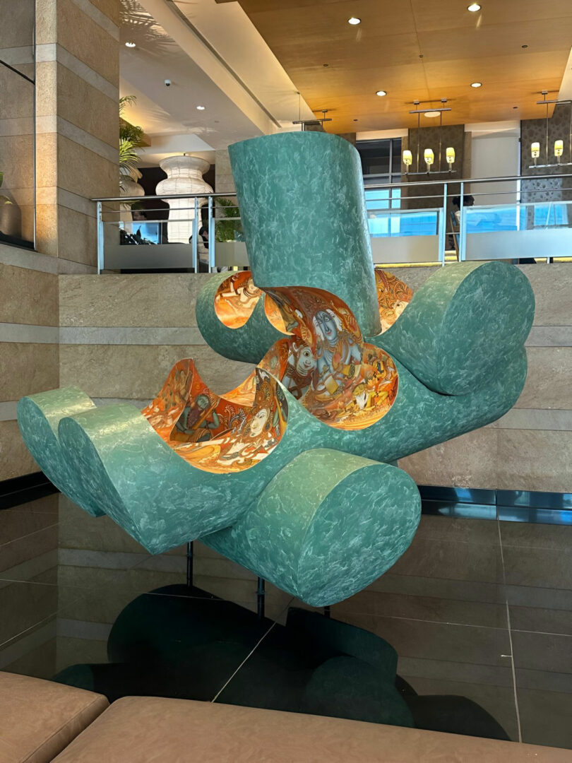 Large abstract green sculpture by Joya Nandurdikar with curved tubular forms, partially hollow and lined with intricate artwork, displayed indoors on a reflective black platform.
