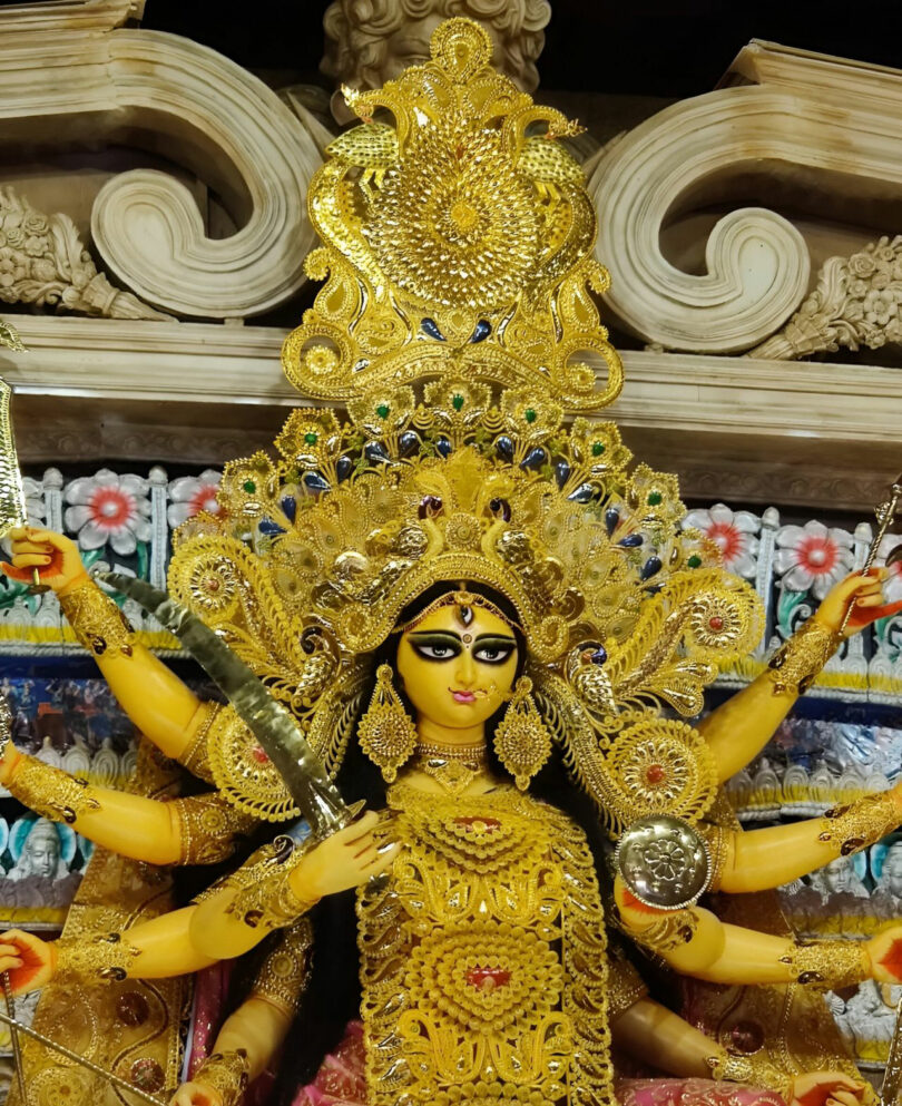 A detailed idol of Goddess Durga adorned with gold ornaments and a crown, holding weapons in her multiple arms, set against a decorative background—an exquisite creation by Joya Nandurdikar.