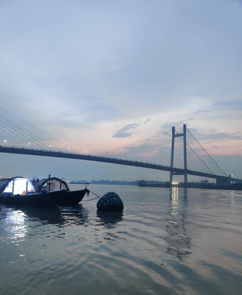 Two boats with lights float on a river at dusk, with a large cable-stayed bridge and city lights in the background—capturing the tranquil evening charm often seen in Joya Nandurdikar's cityscape photography.