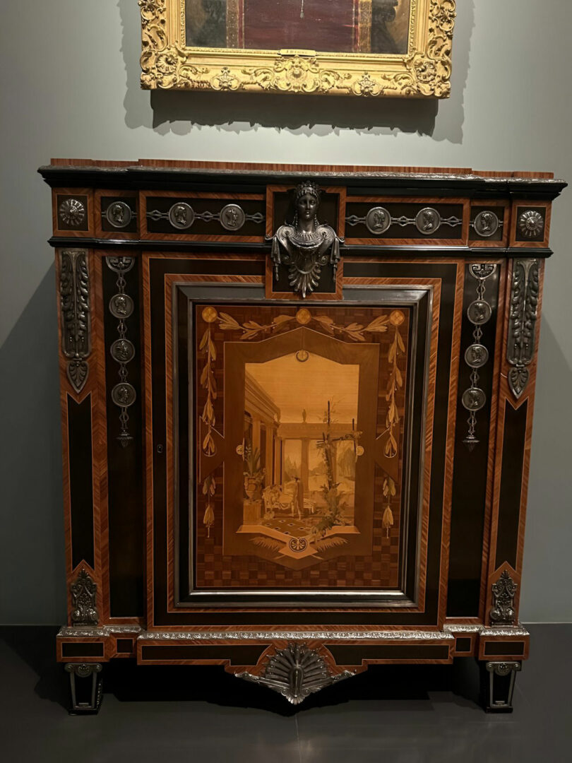 Ornate wooden cabinet with decorative inlays, metal accents, and a central panel depicting a classical architectural scene by Joya Nandurdikar, displayed in a museum setting.