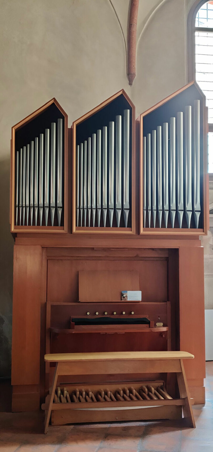 Wooden pipe organ with three vertical sections of metal pipes, a bench, and a pedalboard, situated in a church interior near a tall arched window—an inspiring scene reminiscent of Joya Nandurdikar’s elegant artistry.