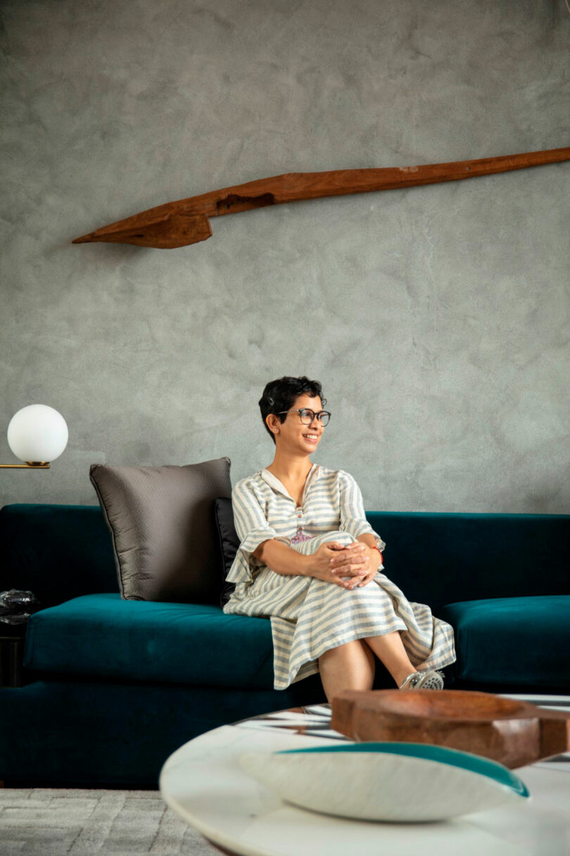 Joya Nandurdikar sits on a teal sofa, wearing a striped dress and glasses, smiling. Behind her is a textured gray wall with a wooden art piece and a side table with a lamp.