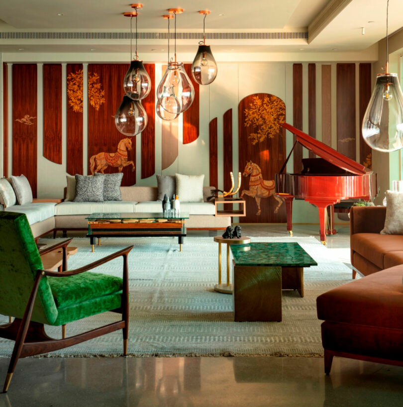 A modern living room with green and brown furniture, a red grand piano, glass pendant lights, and wood paneling featuring Joya Nandurdikar’s signature horse designs on the wall.