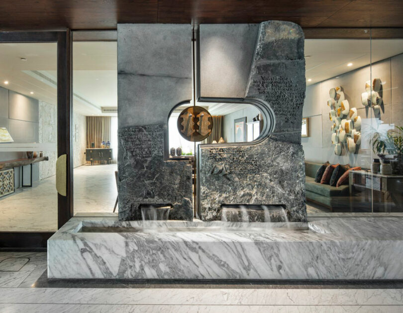 A modern indoor water feature by Joya Nandurdikar, with textured stone, a sculptural circular element, and a marble basin, is situated between two glass-walled living spaces.