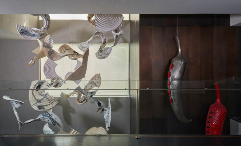 Abstract metallic and ceramic sculptures by Joya Nandurdikar are displayed in a modern indoor setting, with swirling forms on the left and elongated shapes in red and silver on the right, behind a glass barrier.