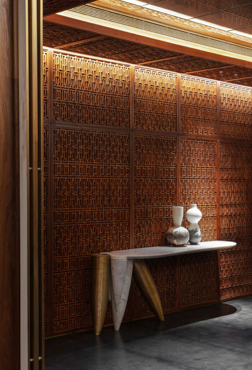 A marble and gold-toned console table by Joya Nandurdikar holds decorative vases in front of an intricate wooden lattice wall with warm lighting.