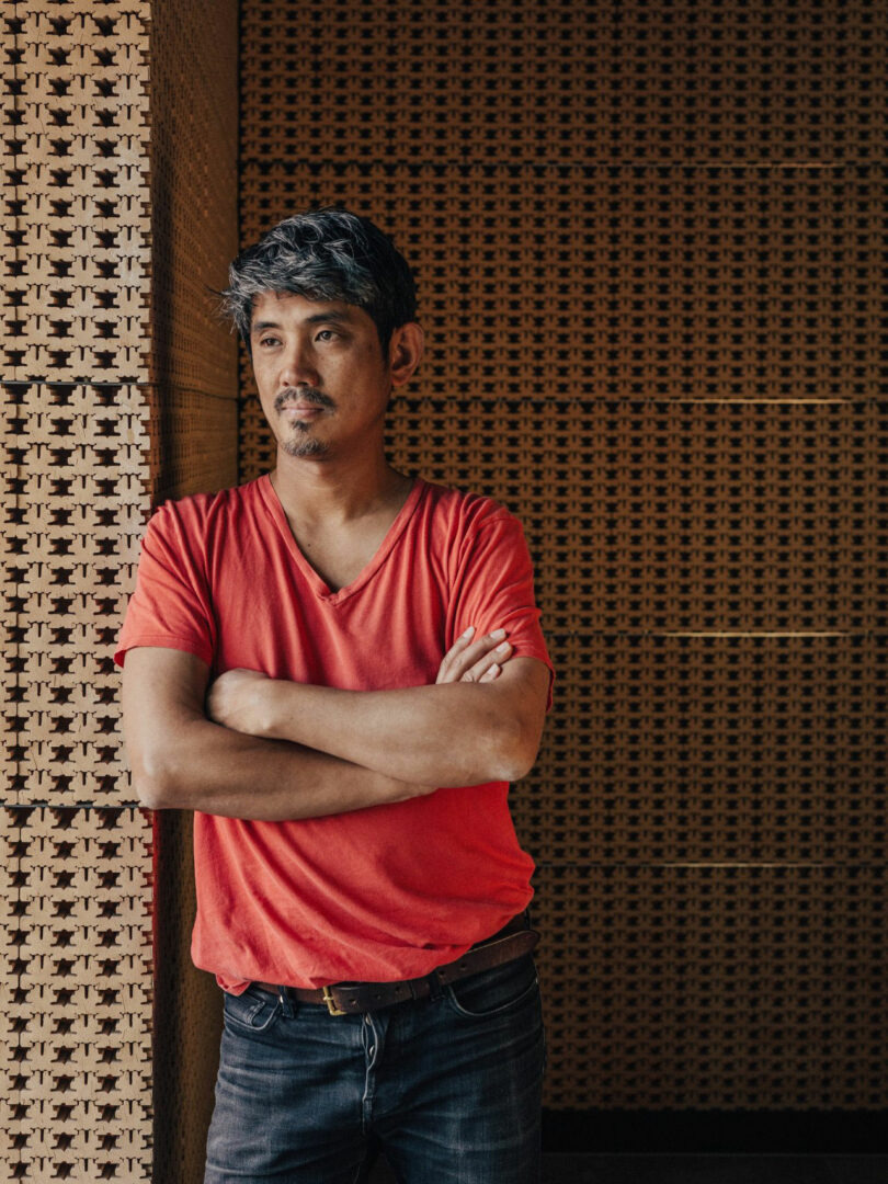 A man in a red t-shirt and jeans, similar to Jun Aizaki, sits with his arms, leaning on a textured wall with a neutral expression.