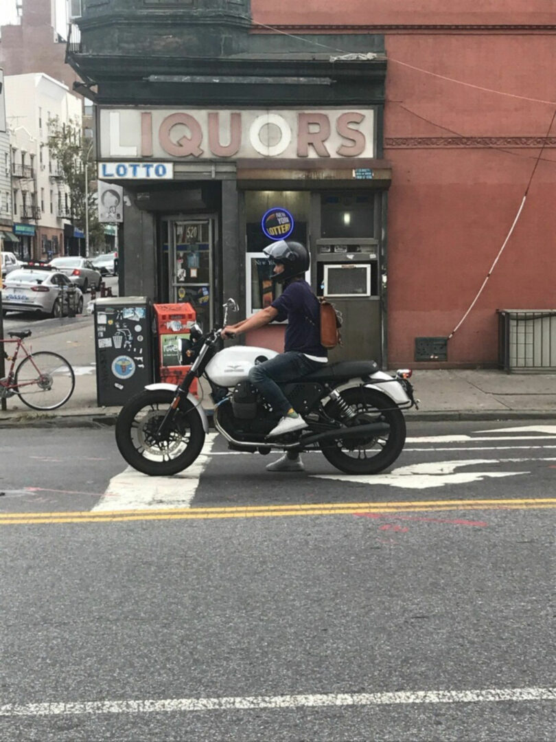 A person wearing helmet and a backpack sits on a white motorcycle stopped on a sidewalk in front of a liqueur store, reminiscent of a scene designed by Jun Aizaki.