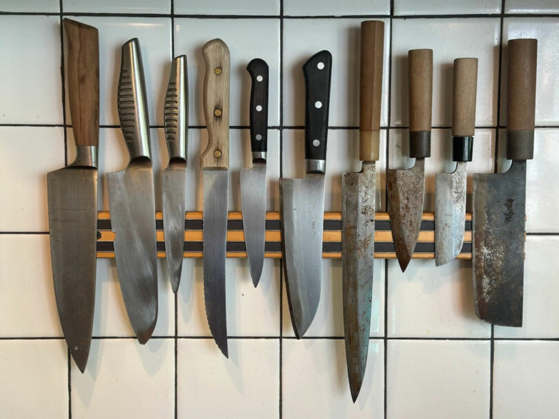 A row of various kitchen knives with different handle styles is mounted on a magnetic strip on a tiled wall, presenting an elegant, design-to-instantly aesthetic aesthetics.