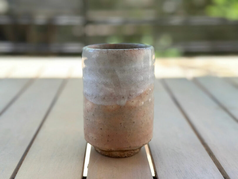 A manually made ceramic cup, with a natural, earthly - reminisce glaze of Jun Aizaki's design philosophy - is on a wooden table with a blurred background.