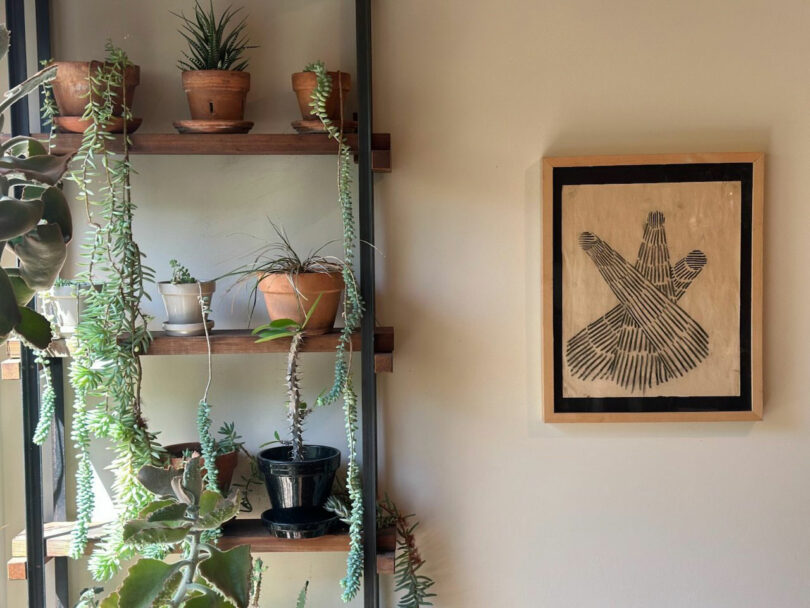 A wooden shelf with plants in the pot is near a wall with a white -white work -abstract black in Jun Aizaki's style.