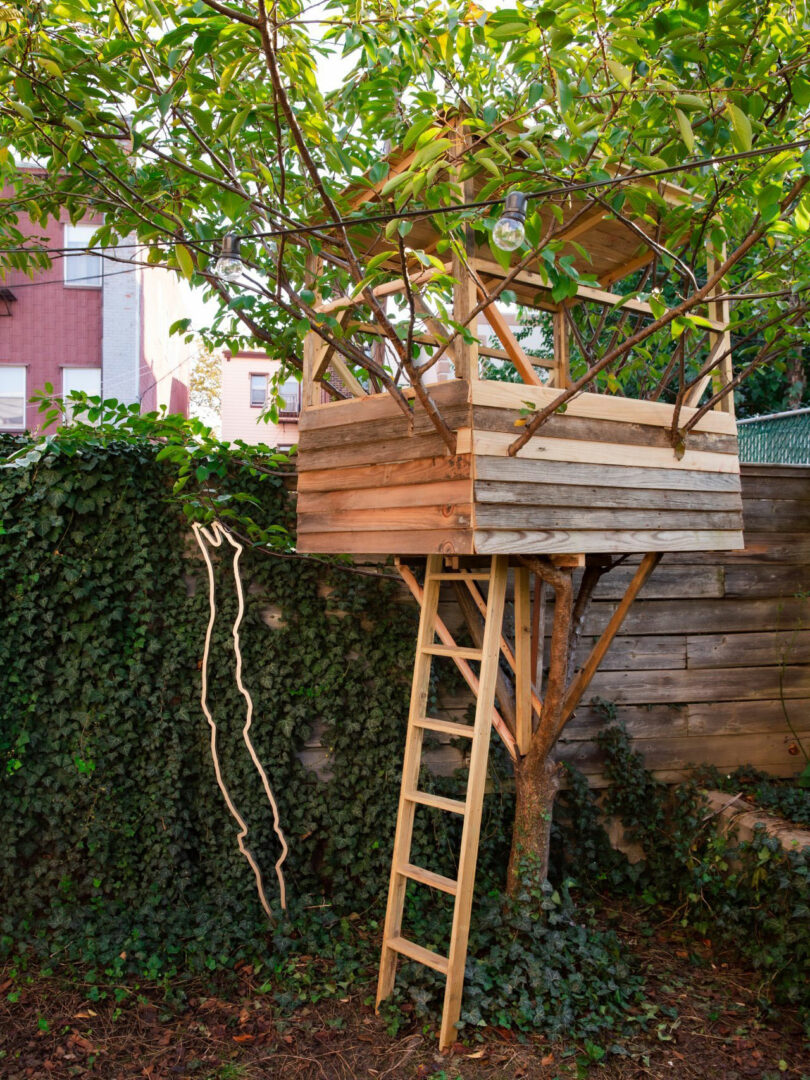 A wooden trees house with a ladder, inspired by Jun Aizaki's natural drawings, is among the branches of a tree in a leaf yard, near a wall covered with vines and wooden fence.