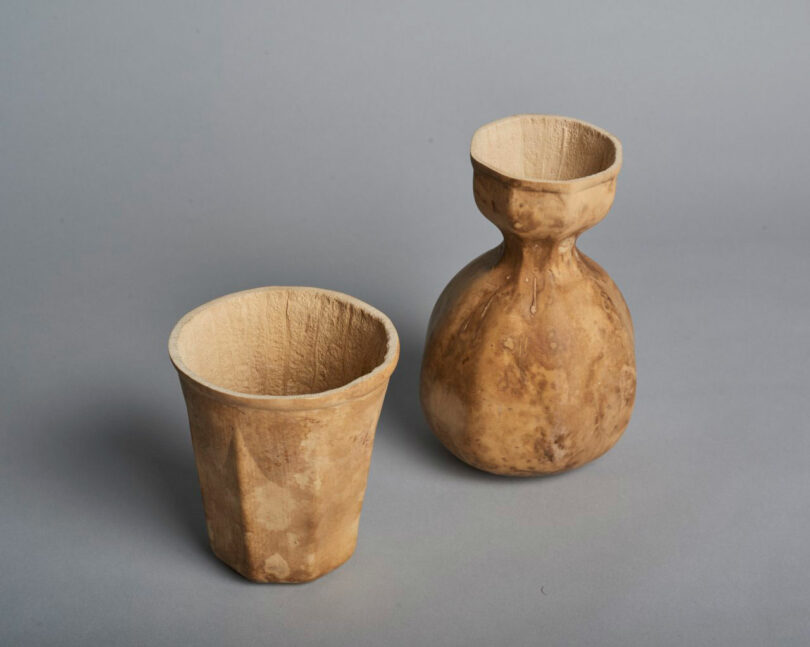 Two brown rustic containers, made of natural-retention material from Jun Aizaki's organic aesthetics-including a right cup and a round-neck, both placed on a simple gray background.