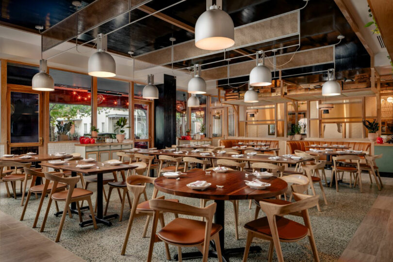 A modern area, well illuminated by the restaurant, designed by Jun Aizaki, with wooden tables and chairs, placed with plates and wipes, and large windows along a part.