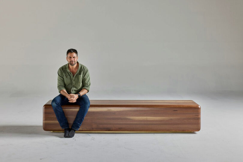 A man in a green shirt and jeans are barefoot on a long bench, rectangular wooden, in a simple, light-colored room.