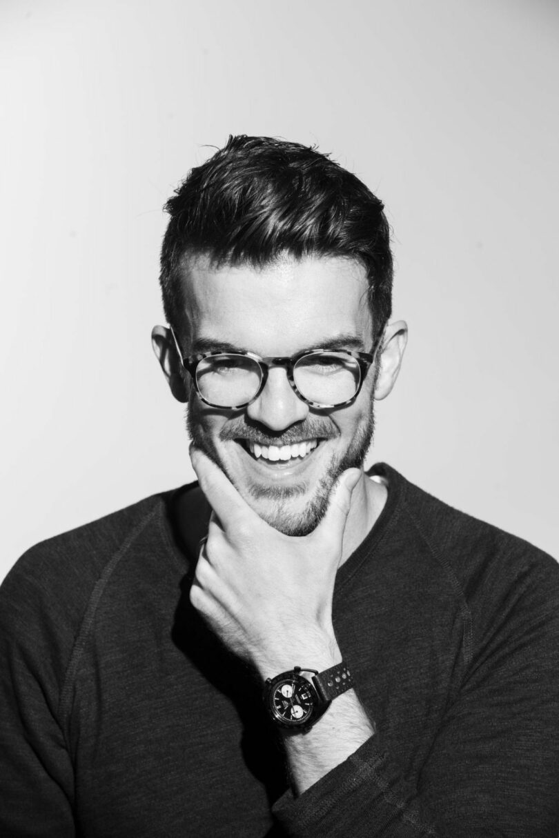 Neil Ferrier, a man with glasses and a beard, smiles while resting his chin on his hand. He is wearing a dark shirt and a wristwatch. The image is in black and white.